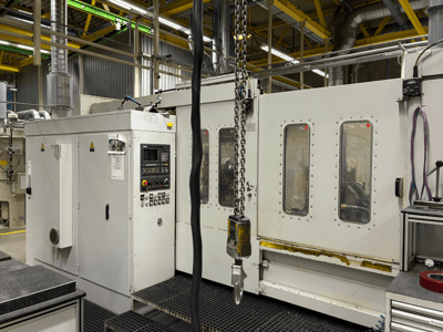 Przemysłowa maszyna CNC z panelem sterowania, widok z boku; zawiera okna, łańcuchy i elementy metalowe w otoczeniu fabrycznym.